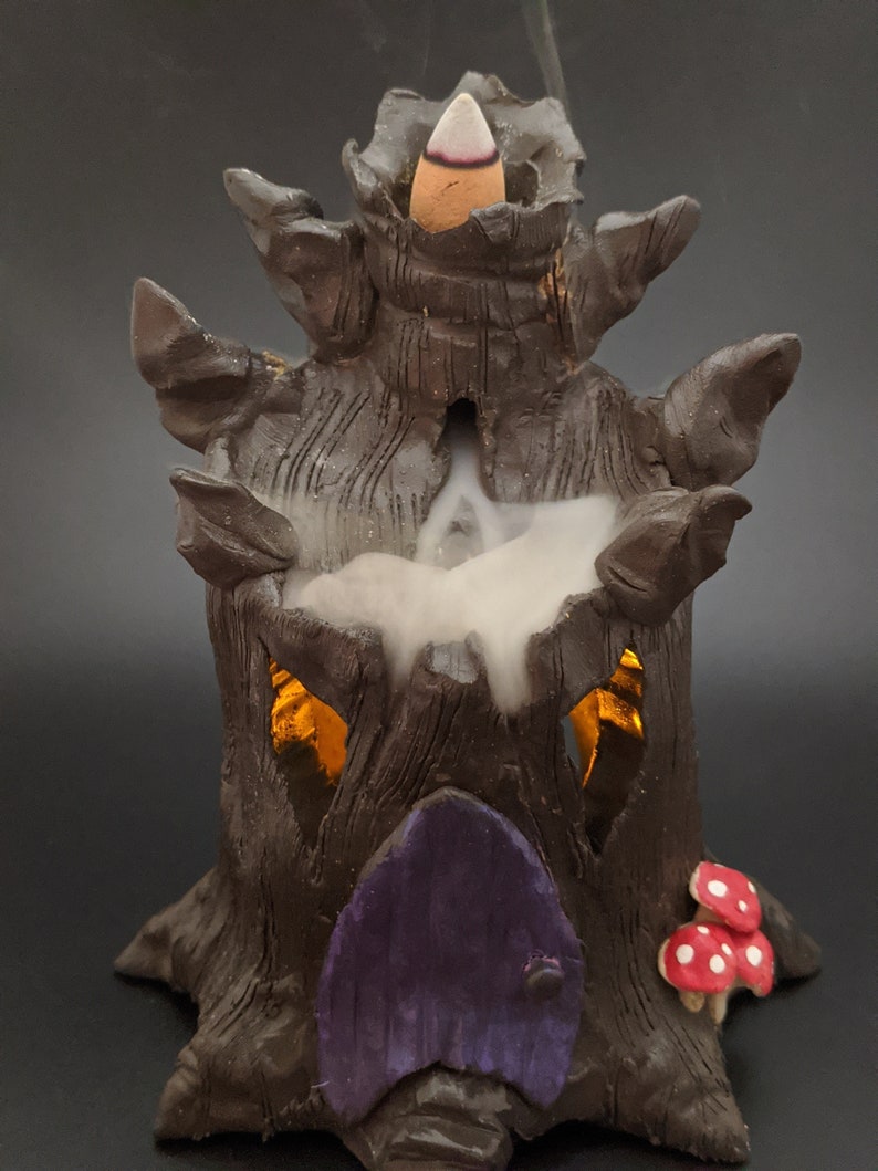 May include: A ceramic incense burner shaped like a tree stump with a purple door and a small red mushroom. Smoke is rising from the top of the burner.