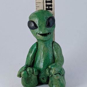 May include: A small green alien figurine with black eyes and a wide smile, sitting with its legs crossed. The figurine is made of clay and has a textured surface.