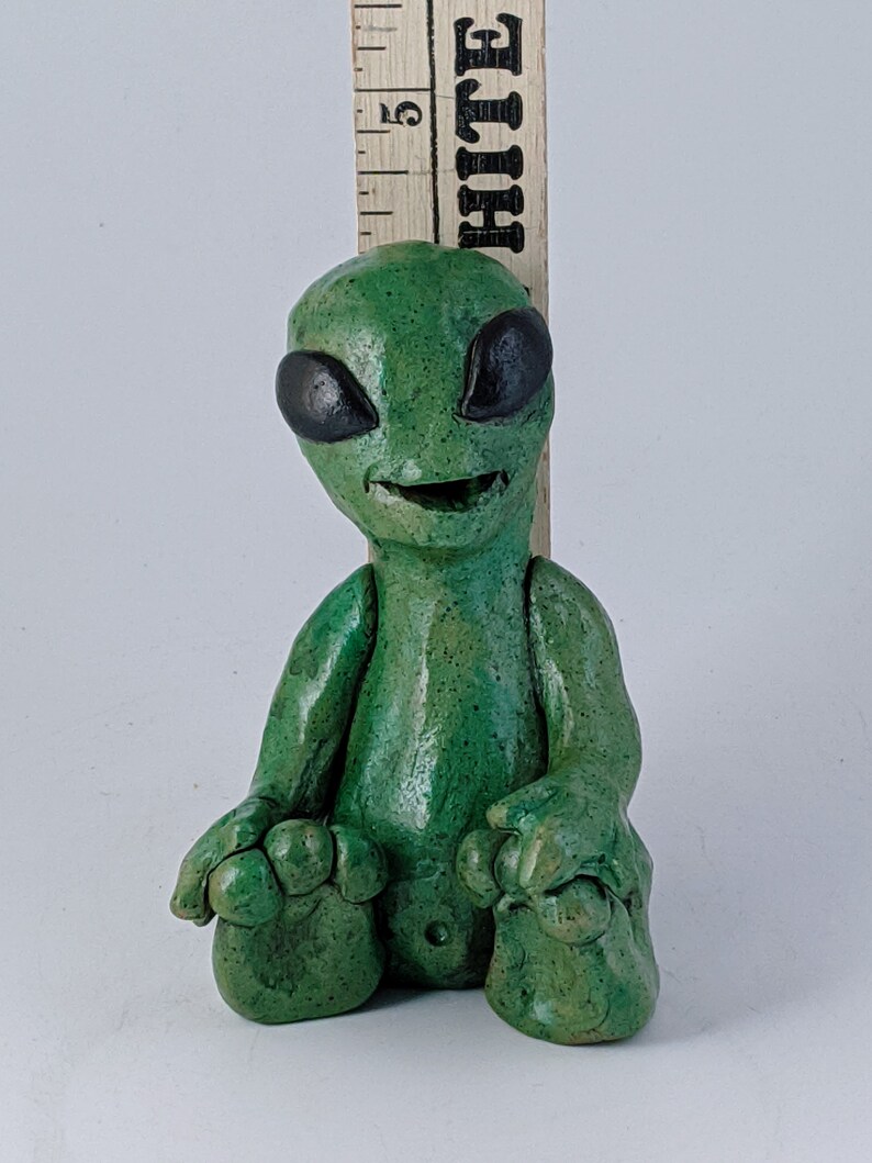 May include: A small green alien figurine with black eyes and a wide smile, sitting with its legs crossed. The figurine is made of clay and has a textured surface.