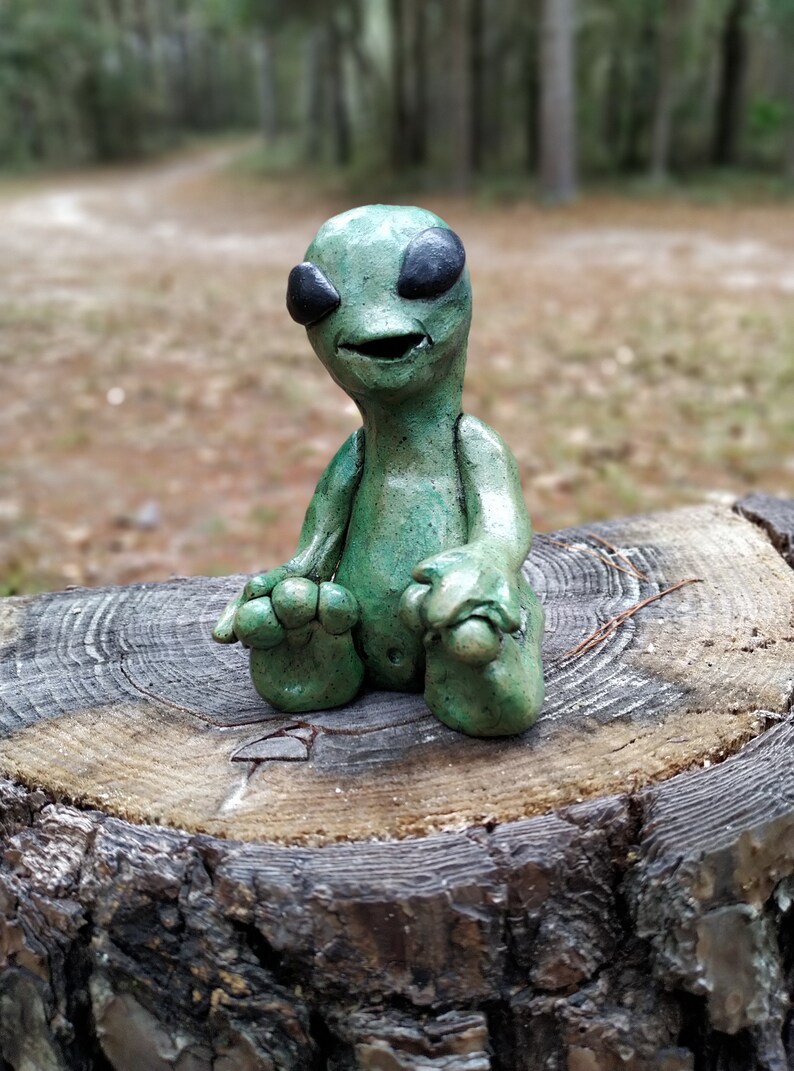May include: A green alien figurine with black eyes and a wide smile, sitting with its legs crossed on a tree stump.