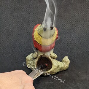 May include: A ceramic incense burner shaped like a gnome with a red, yellow, and green hat. Smoke is rising from the incense burner. The gnome is sitting on a black surface.