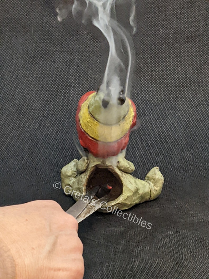 May include: A ceramic incense burner shaped like a gnome with a red, yellow, and green hat. Smoke is rising from the incense burner. The gnome is sitting on a black surface.