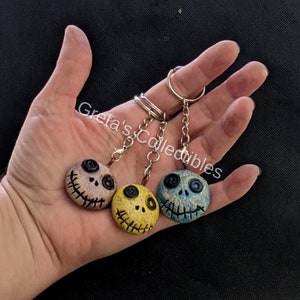 May include: Three keychains with different colored faces. The faces are round and have a stitched mouth, two black eyes, and a black button nose. The keychains are attached to a silver chain and a silver ring.