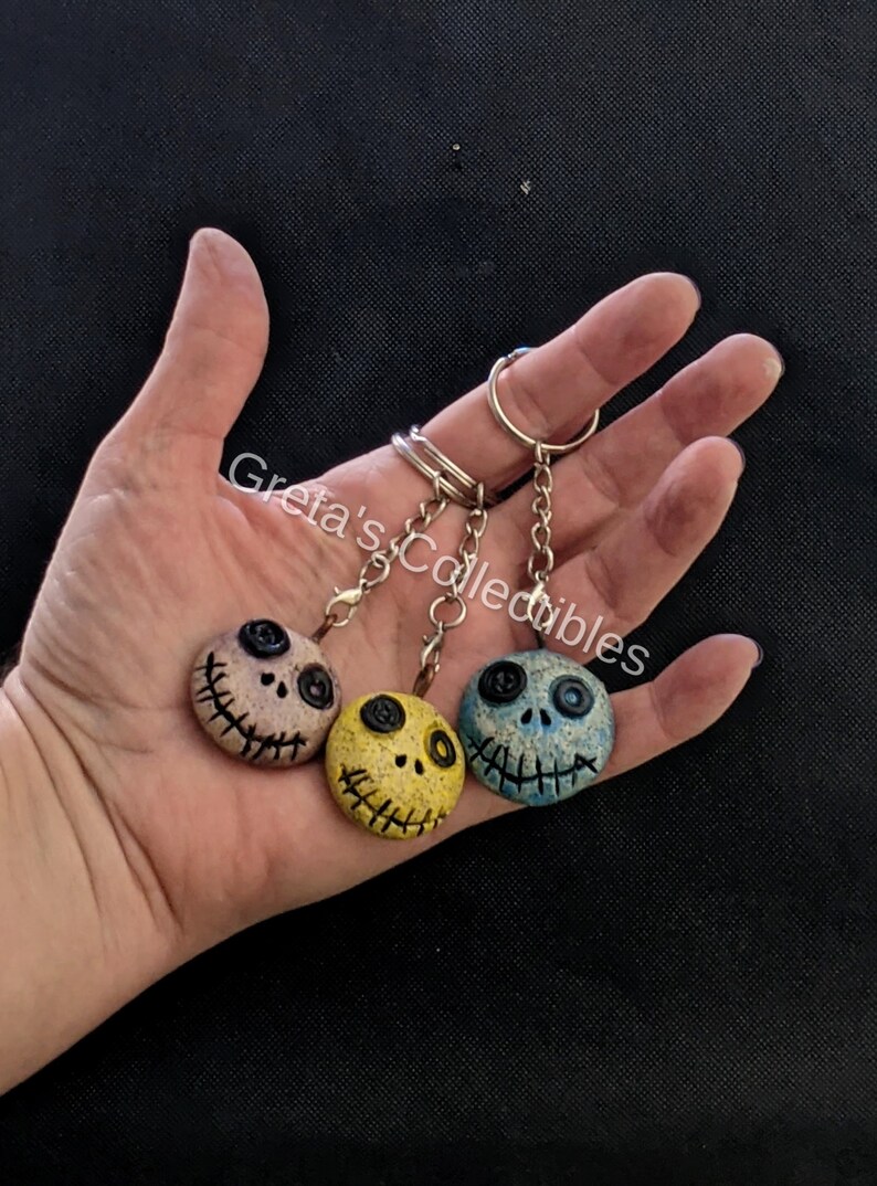 May include: Three keychains with different colored faces. The faces are round and have a stitched mouth, two black eyes, and a black button nose. The keychains are attached to a silver chain and a silver ring.