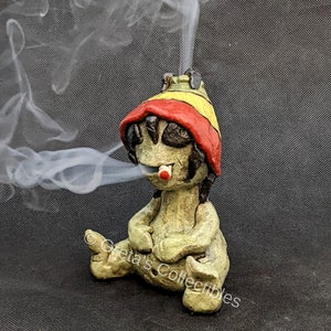 May include: A green ceramic figurine of a person wearing a red and yellow hat, sitting with legs crossed and smoking a cigarette. Smoke is coming from the cigarette.