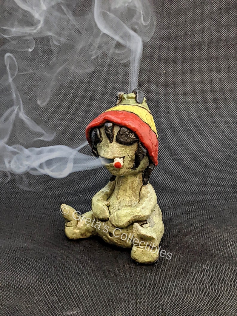 May include: A green ceramic figurine of a person wearing a red and yellow hat, sitting with legs crossed and smoking a cigarette. Smoke is coming from the cigarette.