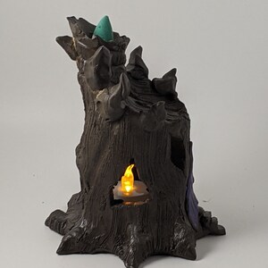 May include: A ceramic incense burner shaped like a tree stump with a teal cone on top and a small candle inside. The tree stump has a dark brown glaze and a purple accent.