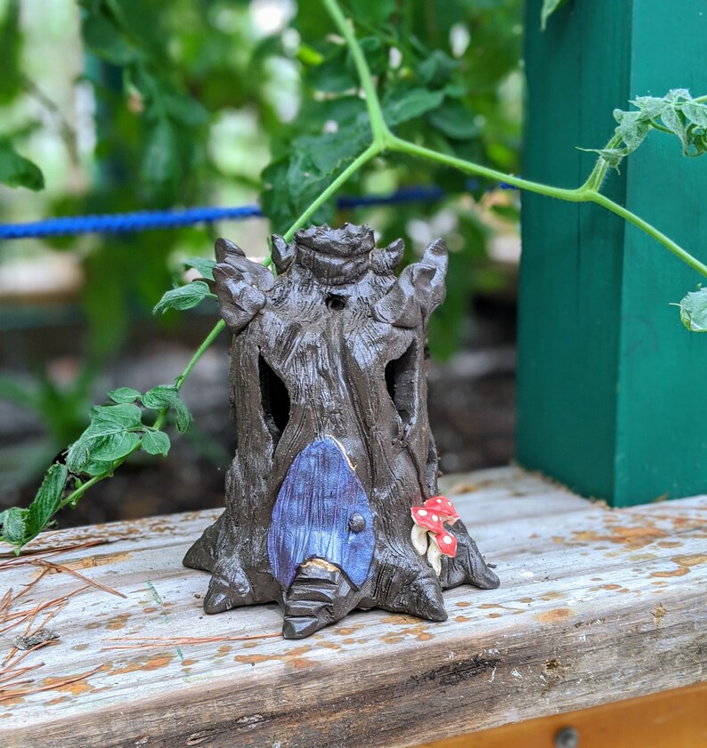 May include: A ceramic treehouse figurine with a blue door and red mushroom accents. The treehouse is made to look like a tree trunk with a hollowed-out space for the door.