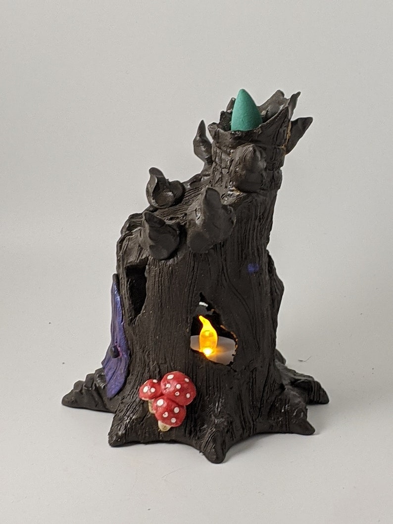 May include: A ceramic incense burner shaped like a tree stump with a teal incense cone in the top, a small candle in a hole, and red and white mushrooms at the base.