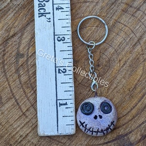 May include: A white and purple clay keychain with a stitched mouth and button eyes. The keychain is attached to a silver key ring and chain.