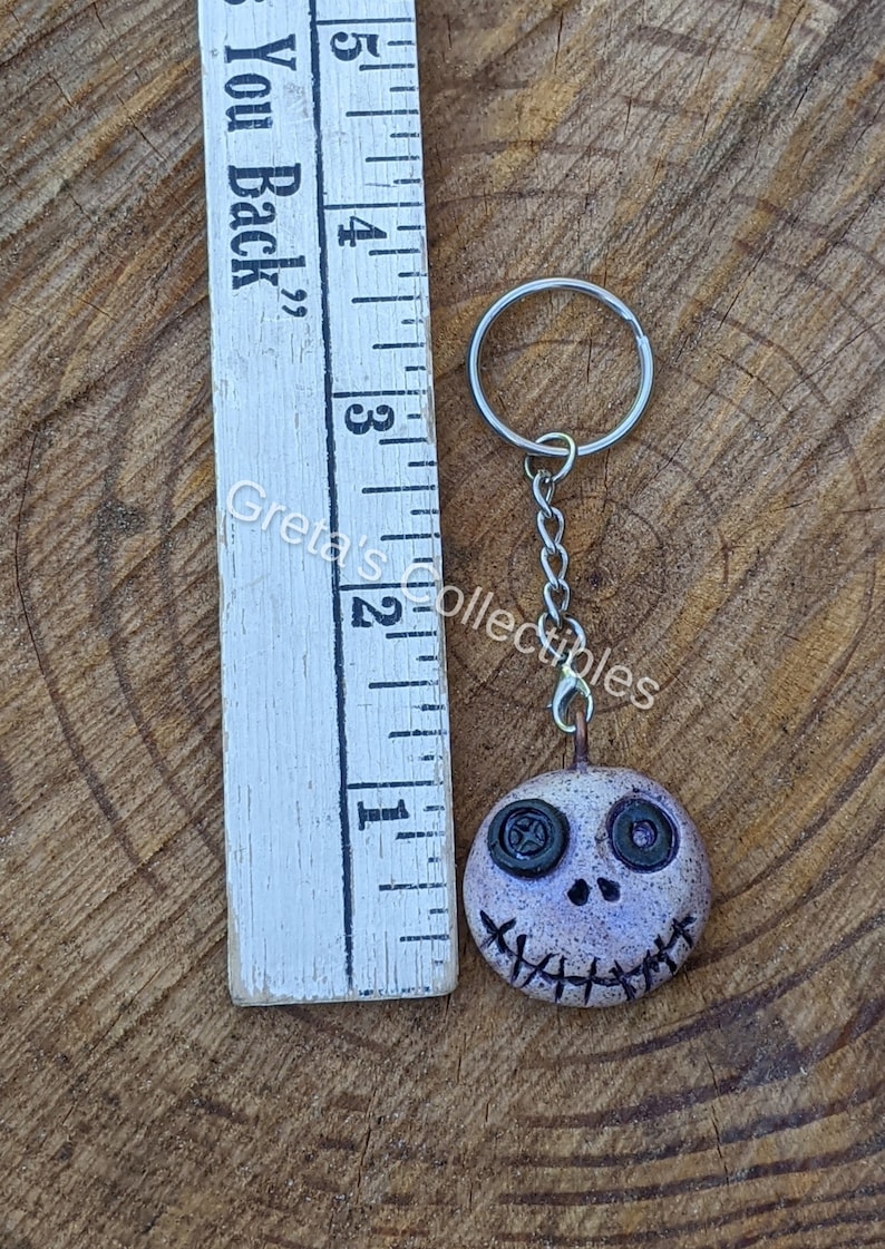 May include: A white and purple clay keychain with a stitched mouth and button eyes. The keychain is attached to a silver key ring and chain.
