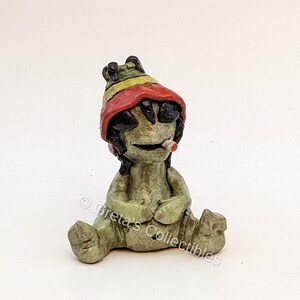 May include: A green ceramic figurine of a sitting figure wearing a red, yellow, and green hat. The figure has black hair and is holding a small white object in its mouth.