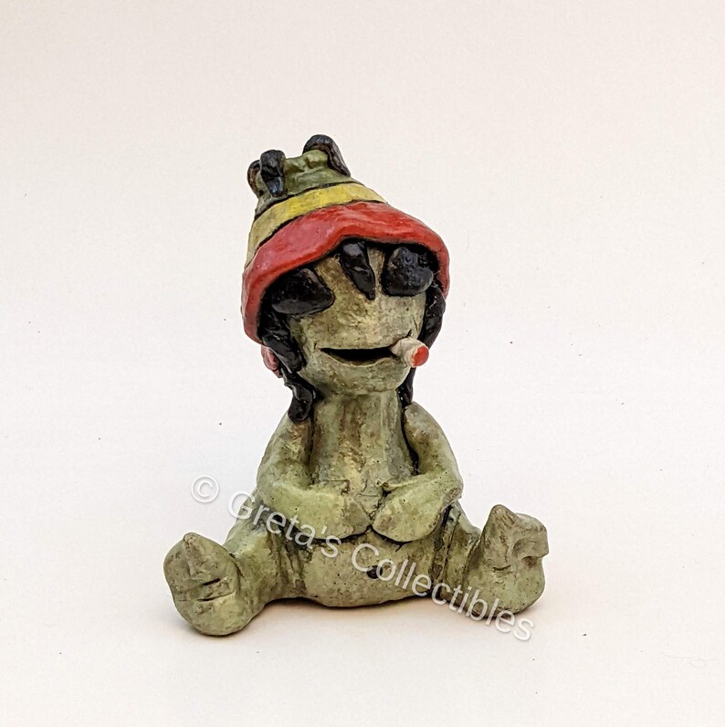 May include: A green ceramic figurine of a sitting figure wearing a red, yellow, and green hat. The figure has black hair and is holding a small white object in its mouth.