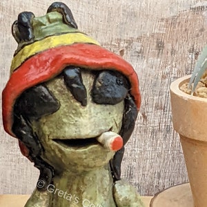 May include: A green ceramic figurine of a person wearing a red, yellow, and green hat and holding a red cigarette. The figurine has a black face and black hair.