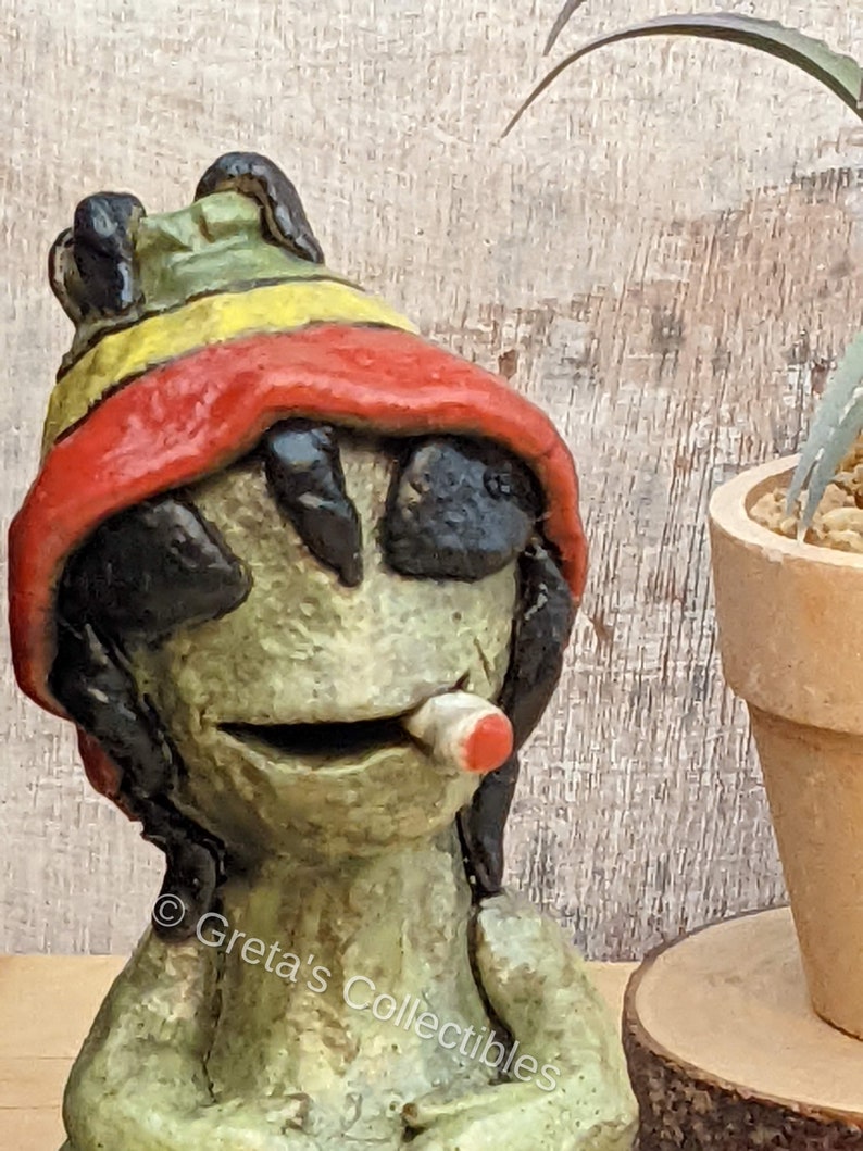 May include: A green ceramic figurine of a person wearing a red, yellow, and green hat and holding a red cigarette. The figurine has a black face and black hair.