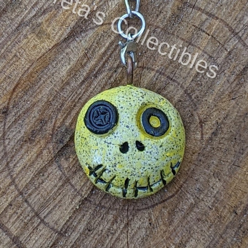 May include: A yellow and black skull charm with a stitched mouth and button eyes. The charm is attached to a silver key chain.