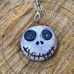 May include: A white ceramic charm with a stitched mouth, button eyes, and a silver chain. The charm is shaped like a face and has a spooky, whimsical look.