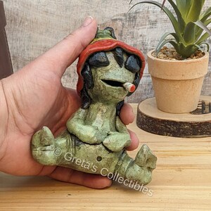 May include: A green ceramic figurine of a person sitting with legs crossed, wearing a red, yellow, and green hat. The figurine has a small white pipe in its mouth. The figurine is sitting on a wooden surface.