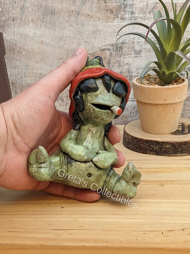 May include: A green ceramic figurine of a person sitting with legs crossed, wearing a red, yellow, and green hat. The figurine has a small white pipe in its mouth. The figurine is sitting on a wooden surface.