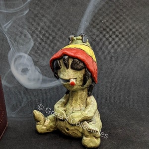 May include: A green ceramic figurine of a person wearing a red and yellow hat, sitting with legs crossed and smoking a cigarette. The figurine is a incense burner with smoke coming out of the top of the hat.