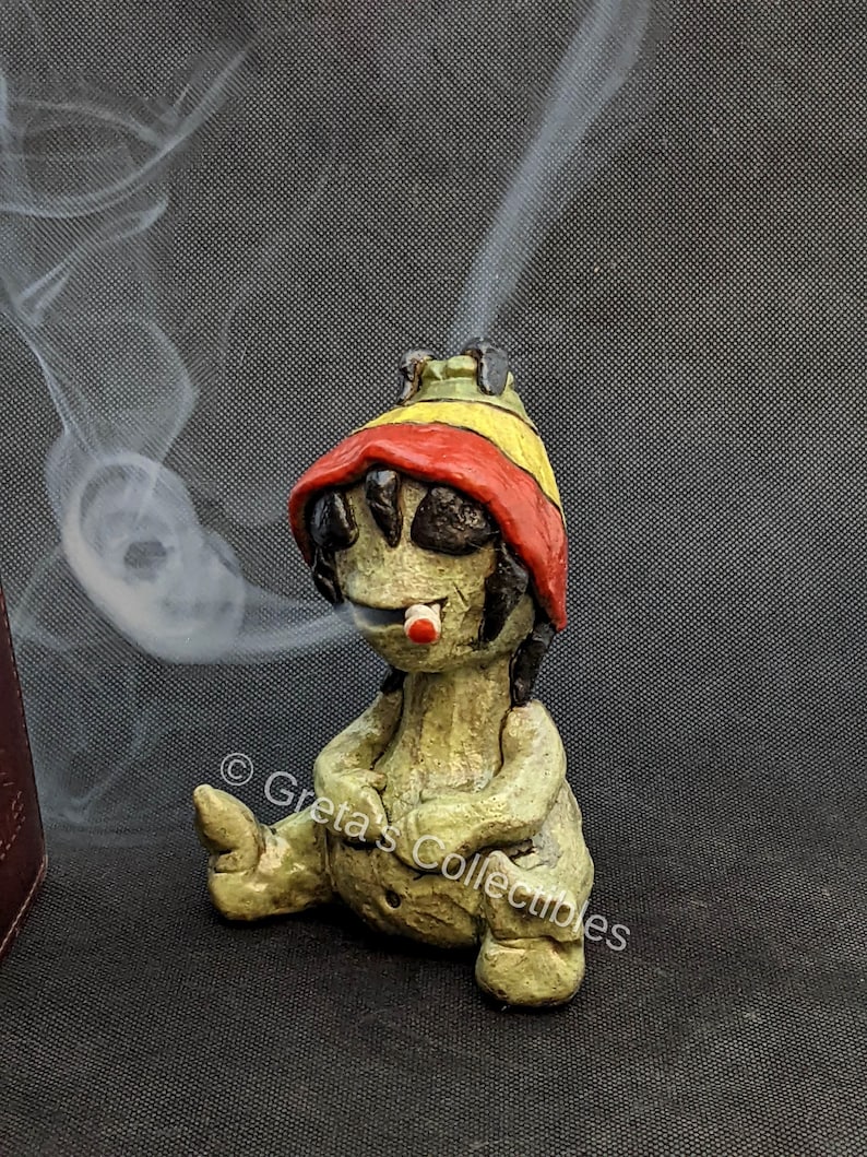 May include: A green ceramic figurine of a person wearing a red and yellow hat, sitting with legs crossed and smoking a cigarette. The figurine is a incense burner with smoke coming out of the top of the hat.