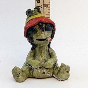 May include: A green ceramic figurine of a sitting figure with a red, yellow, and green hat, wearing sunglasses and holding a cigarette. The figurine is approximately 5 inches tall.