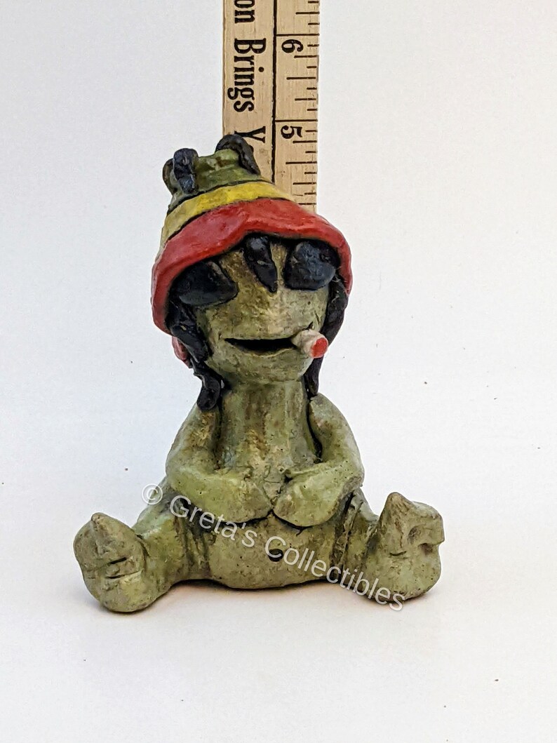 May include: A green ceramic figurine of a sitting figure with a red, yellow, and green hat, wearing sunglasses and holding a cigarette. The figurine is approximately 5 inches tall.