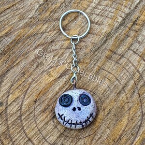 May include: A white and gray clay keychain with a stitched mouth and black button eyes. The keychain is attached to a silver chain and ring.