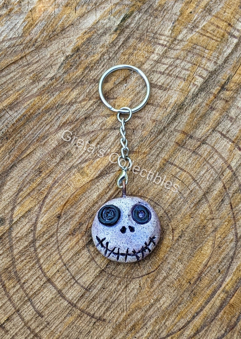 May include: A white and gray clay keychain with a stitched mouth and black button eyes. The keychain is attached to a silver chain and ring.