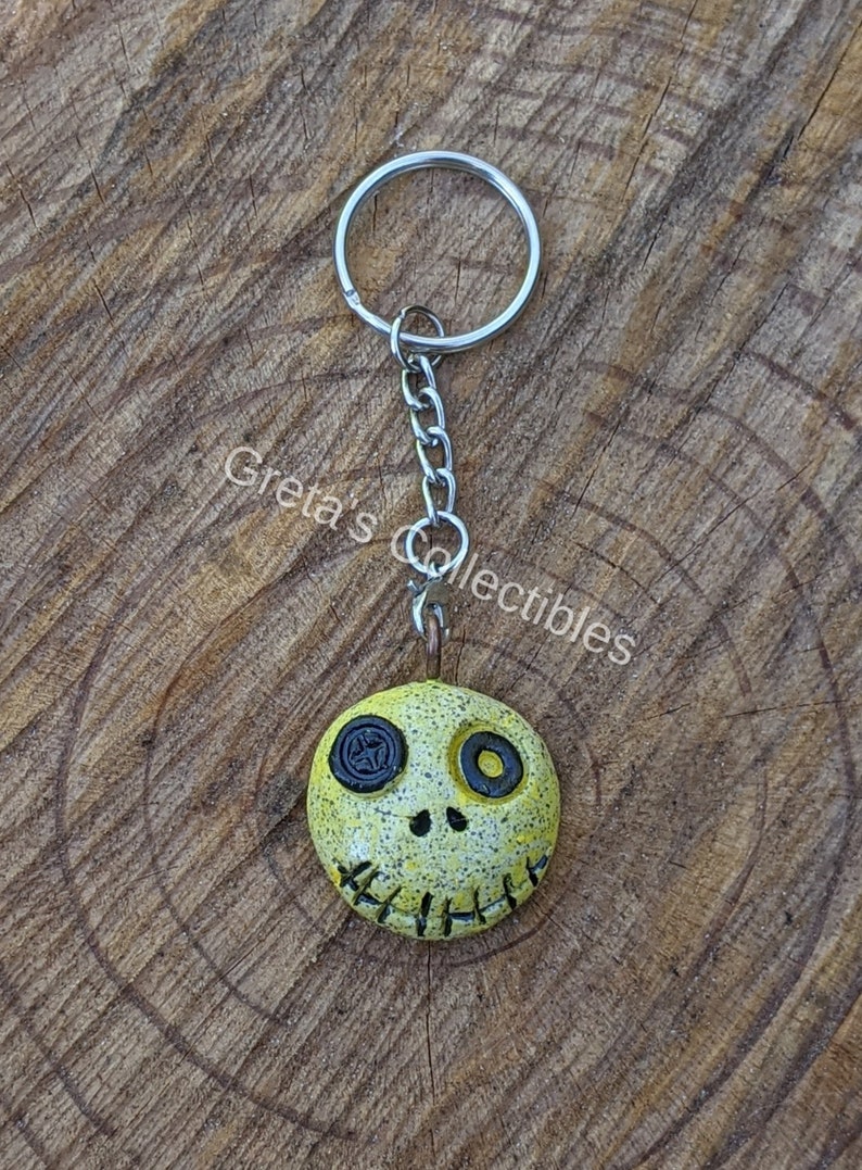 May include: Yellow and black clay charm with a stitched mouth and button eyes on a silver key chain.
