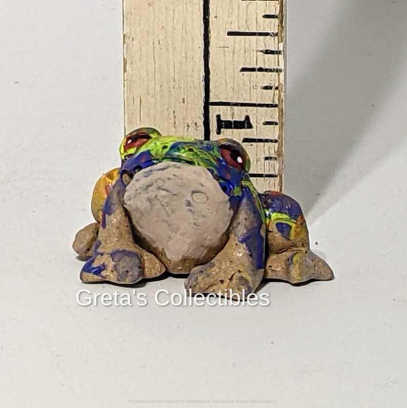 May include: A small, hand-painted ceramic frog figurine with a blue, green, yellow, and red paint design. The frog is sitting on its belly with its legs tucked underneath it. The frog is about 2 inches tall.