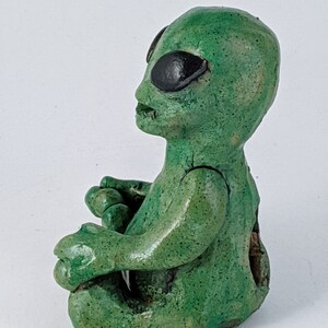 May include: Green ceramic alien figurine sitting in a meditative pose with its legs crossed. The alien has large black eyes and a textured surface.