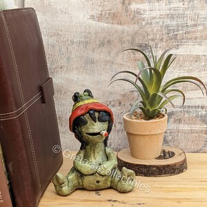 May include: A green ceramic figurine of a person wearing a red, yellow, and green hat and sunglasses, sitting with their legs crossed and holding a cigarette. The figurine is sitting on a wooden surface next to a potted plant.