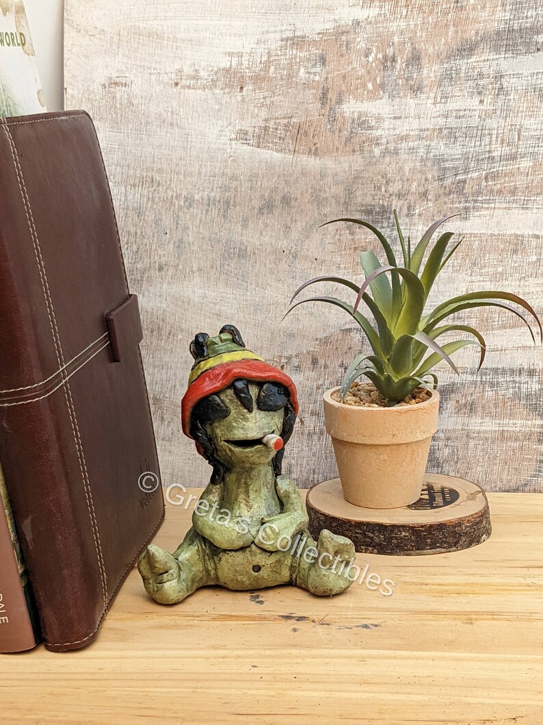 May include: A green ceramic figurine of a person wearing a red, yellow, and green hat and sunglasses, sitting with their legs crossed and holding a cigarette. The figurine is sitting on a wooden surface next to a potted plant.