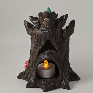 May include: A brown ceramic tree stump incense burner with a teal green top and a small red mushroom at the base. A white tea light candle is placed inside the hollow of the stump, illuminating the interior.