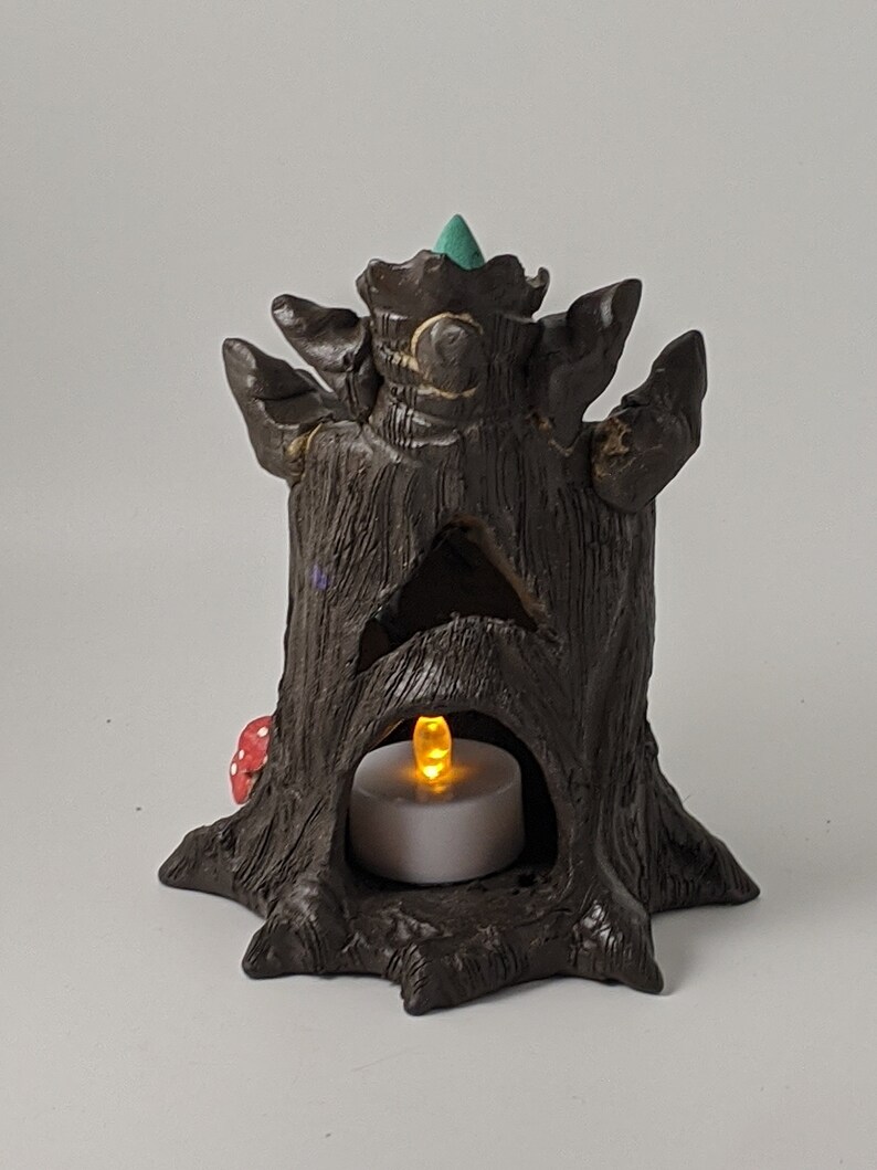 May include: A brown ceramic tree stump incense burner with a teal green top and a small red mushroom at the base. A white tea light candle is placed inside the hollow of the stump, illuminating the interior.