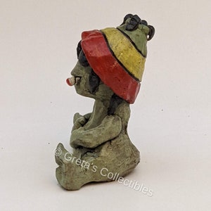 May include: A small ceramic figurine of a seated figure wearing a red, yellow, and green hat. The figure has a green body and is holding a small object in its hands. The figurine is sitting on a white surface. &copy; Greta's Collectibles