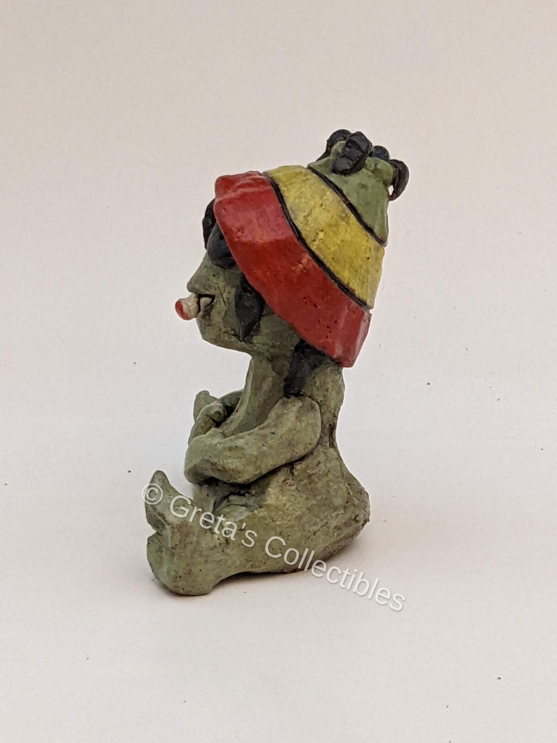 May include: A small ceramic figurine of a seated figure wearing a red, yellow, and green hat. The figure has a green body and is holding a small object in its hands. The figurine is sitting on a white surface. &copy; Greta's Collectibles