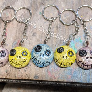 May include: Six colorful, hand-painted, ceramic keychains with a stitched-mouth skull design. Each keychain features a different color scheme, including yellow, blue, and pink.