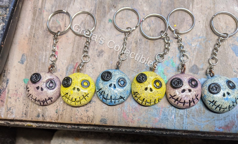 May include: Six colorful, hand-painted, ceramic keychains with a stitched-mouth skull design. Each keychain features a different color scheme, including yellow, blue, and pink.