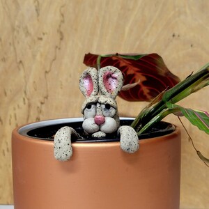 May include: A ceramic bunny rabbit figurine with pink ears and a brown pot with green plants. The bunny is peeking out of the pot.