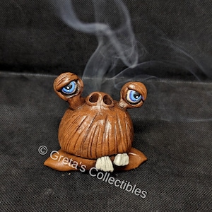 May include: A brown ceramic incense burner shaped like a creature with two large eyes, a wide mouth with white teeth, and a textured surface. Smoke is rising from the top of the burner.