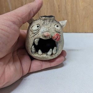May include: A small, white ceramic figurine of a monster with a wide open mouth, large eyes, and a tongue sticking out. The monster has a textured surface and a black rim around the top of its head.