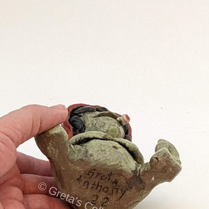 May include: A green ceramic figurine of a gnome with a red hat and a white beard. The gnome is sitting with its legs crossed and its hands on its knees. The figurine is signed "Greta Anthony 22" on the bottom.