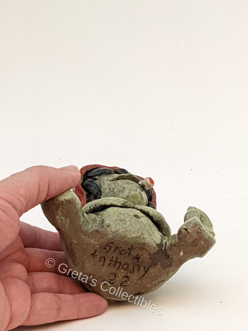 May include: A green ceramic figurine of a gnome with a red hat and a white beard. The gnome is sitting with its legs crossed and its hands on its knees. The figurine is signed "Greta Anthony 22" on the bottom.