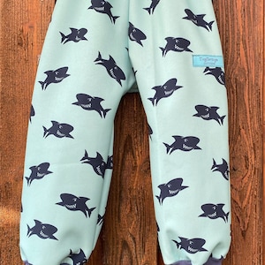 Könnte beinhalten: Hellblaue Kinderhose mit Hai-Muster. Die Hose hat einen dunkelblauen Bund und Bündchen. Die Hose hat ein kleines Etikett mit der Aufschrift "TinyTrousers Outdoor".