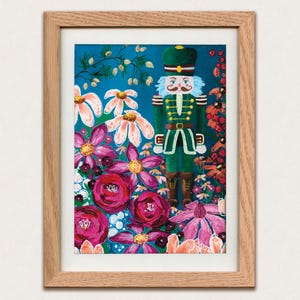 Puede incluir: Una impresión artística enmarcada que presenta un cascanueces rodeado de flores vibrantes. El cascanueces, con uniforme verde y detalles dorados, se alza sobre un fondo turquesa. La obra está enmarcada en un marco de madera marrón claro.