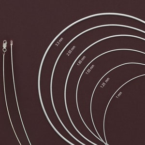 May include: A set of seven silver snake chains of varying widths, ranging from 1 mm to 3.3 mm. The chains are arranged in a circular pattern on a dark brown background.