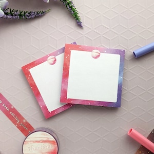 Può includere: Due foglietti adesivi quadrati con un bordo a tema galassia nei toni del rosso, rosa e blu. Ogni foglietto ha un piccolo adesivo a forma di pianeta in alto. Sono visibili anche un rotolo di nastro washi abbinato e una penna viola.