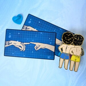 May include: Gold enamel pin featuring two figures with black hair, wearing blue and yellow swim trunks, standing back to back with their arms around each other. The figures are inspired by the famous "Creation of Adam" painting. The pin is on a blue background with a grid pattern.